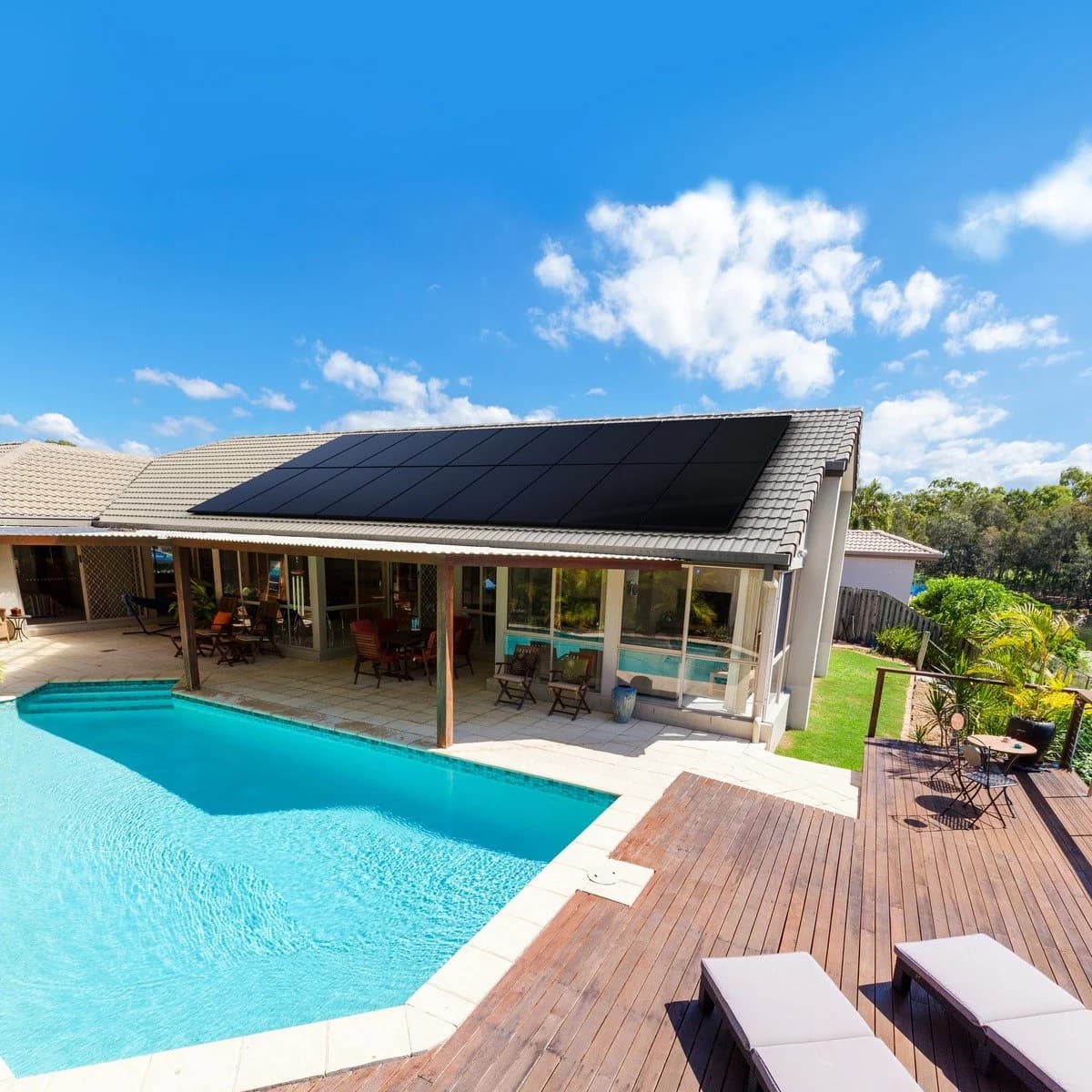 Modern single-story house with tiled roof featuring a large array of sleek all-black solar panels, overlooking a swimming pool and outdoor patio area on a sunny day with blue sky.