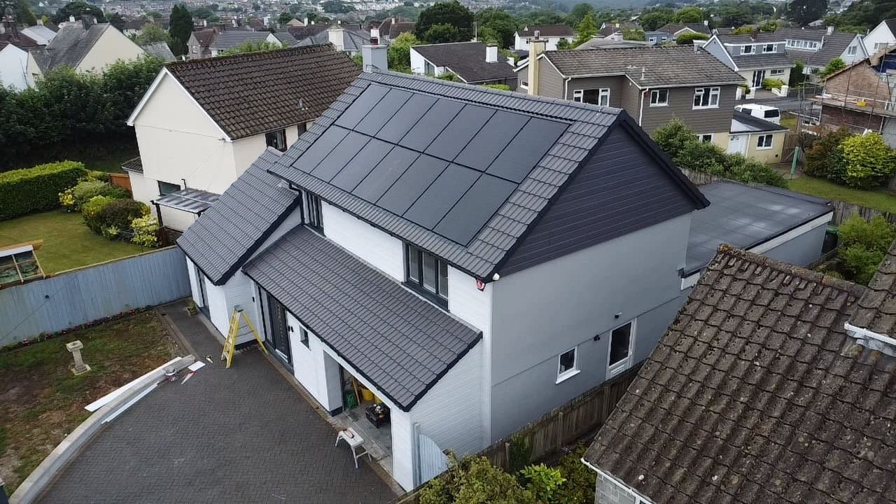 An aerial view of a residential house with solar panels on the roof, neighboring houses, a backyard with a garden, and a fenced yard.