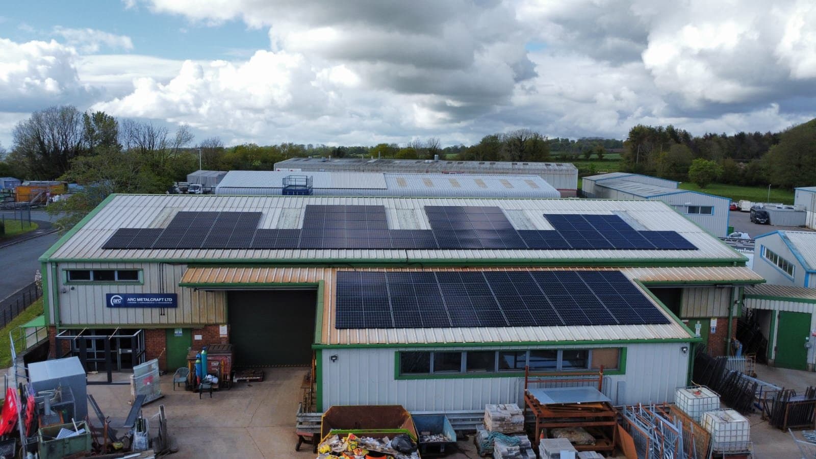 Industrial warehouse buildings with solar panels on the roofs, surrounded by outdoor storage and greenery under a cloudy sky.
