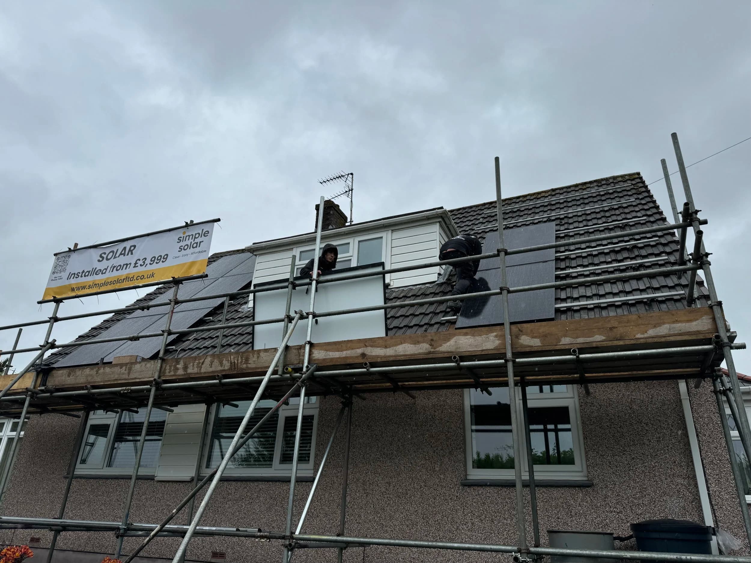 Solar installation in progress on a house roof with scaffolding and a Simple Solar banner, under an overcast sky.