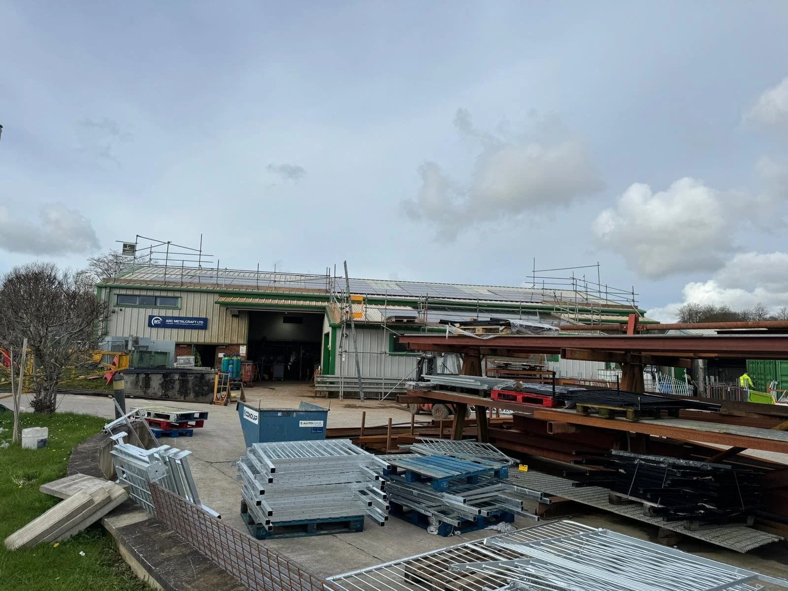 Industrial workshop yard with scaffolding on the roof and stacks of steel beams and metal panels in the foreground.