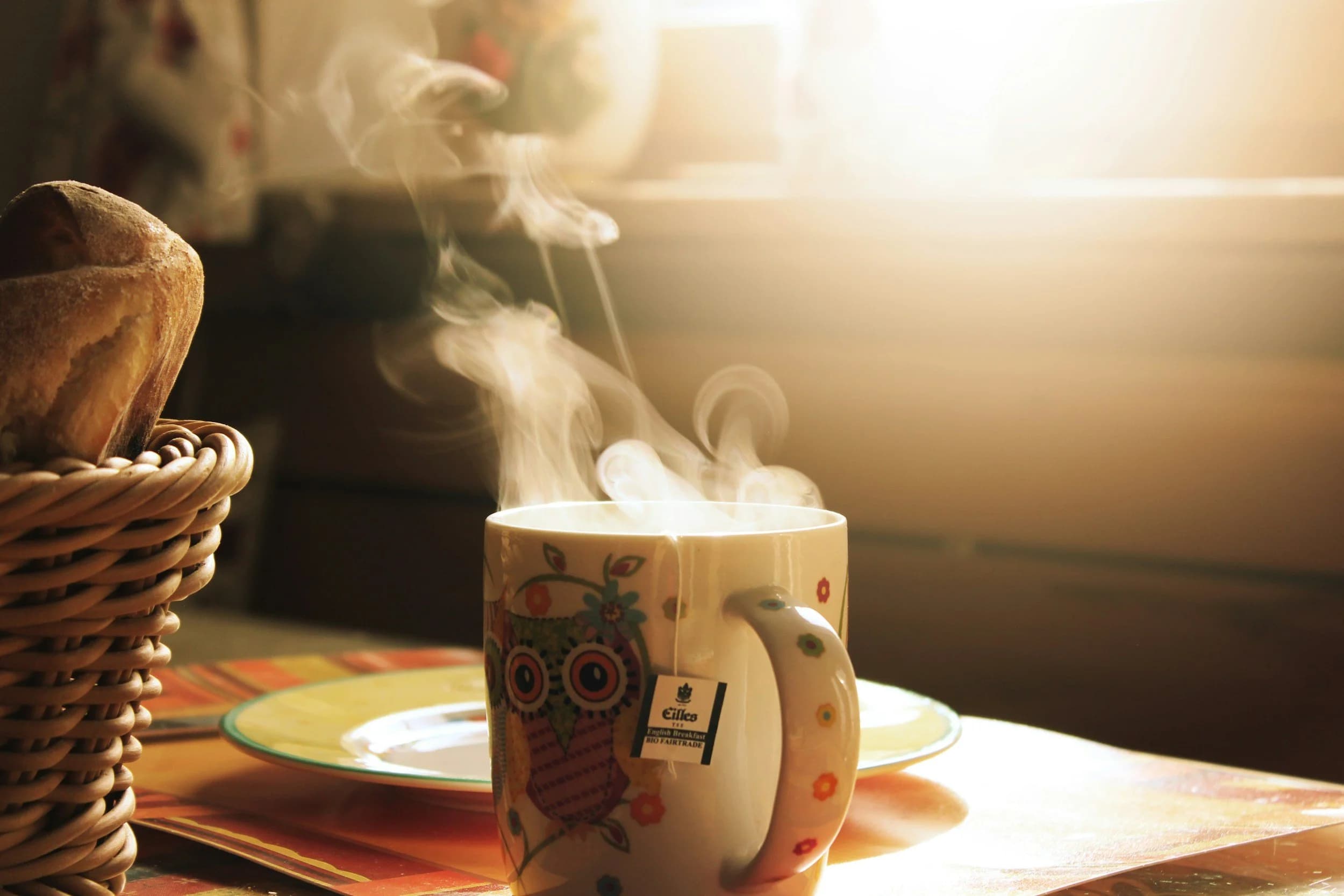 Steaming hot cup of tea in a colorful owl-patterned mug with an Eilles English Breakfast Fairtrade tea bag tag, placed on a saucer next to a wicker basket of bread rolls on a cozy table.