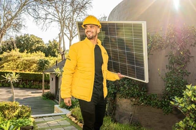 A man in a yellow jacket and hard hat holding a solar panel outdoors on a sunny day.
