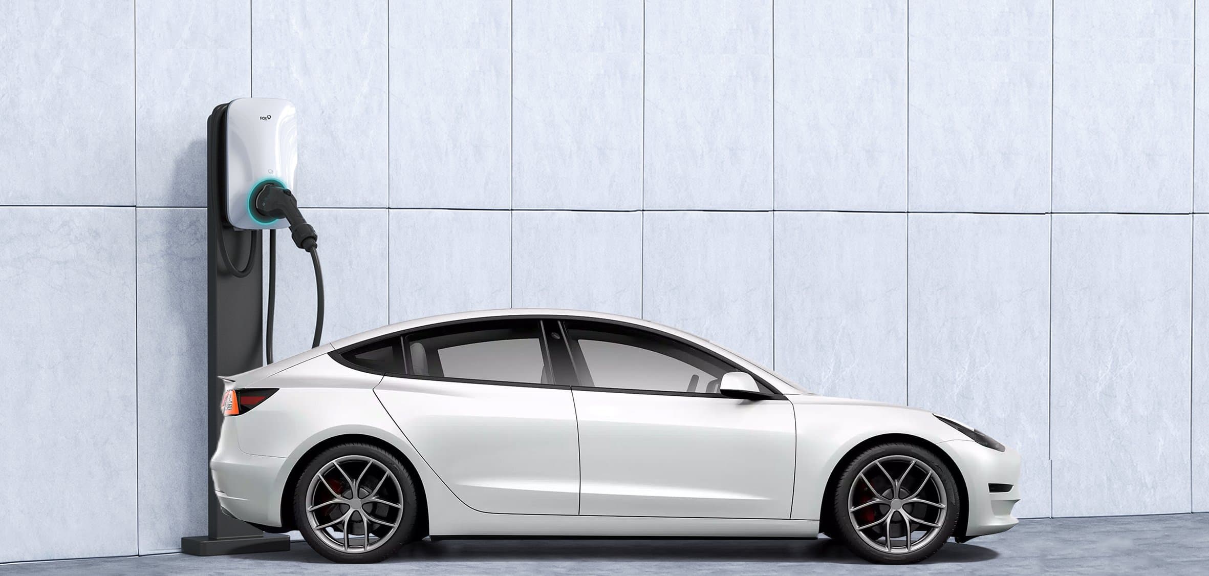 White Tesla Model 3 parked indoors and connected to a wall-mounted Fox ESS EV charger with a teal accent, against a light gray tiled wall.