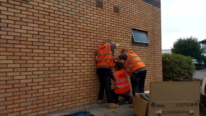 Three workers in orange “NET” hi-vis vests pressed against a brick wall corner, seemingly hiding; one kneels. Cardboard box labeled “ATESS DC CHARGING PILE” in foreground.